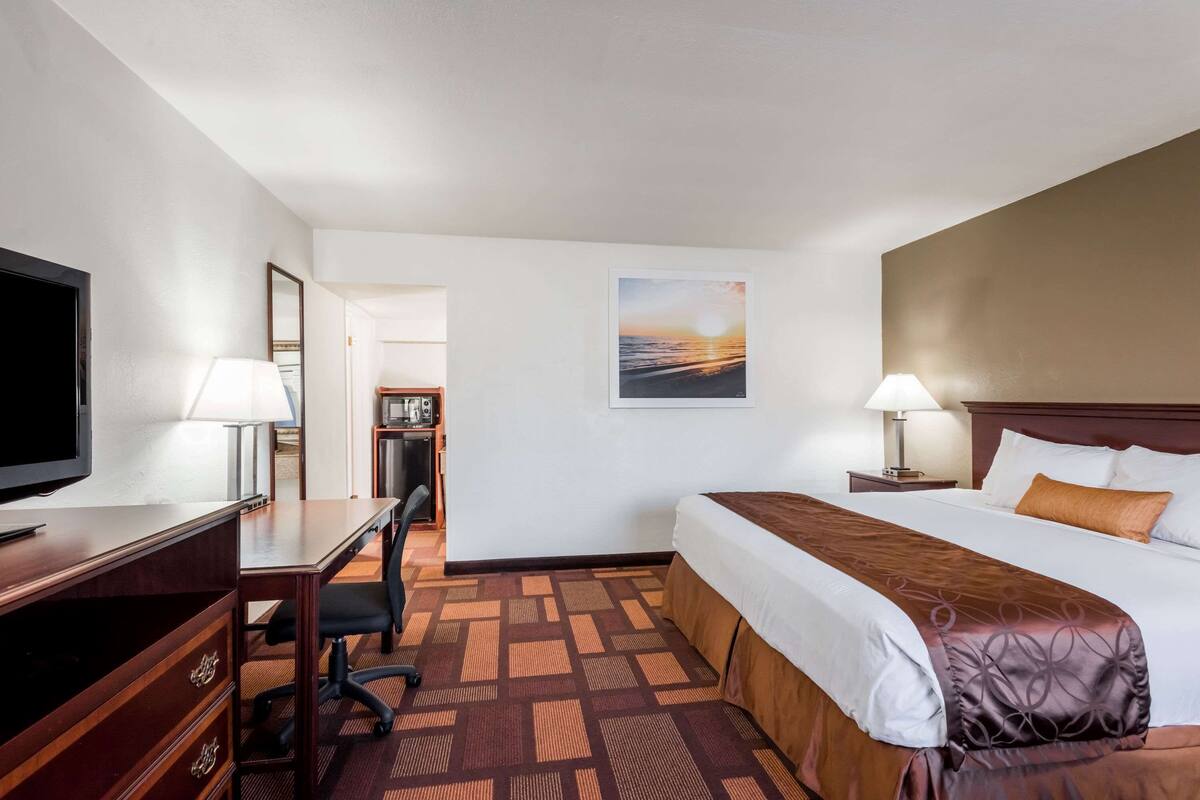 A hotel room with a bed, desk, chair, TV, lamps, and a painting on the wall.