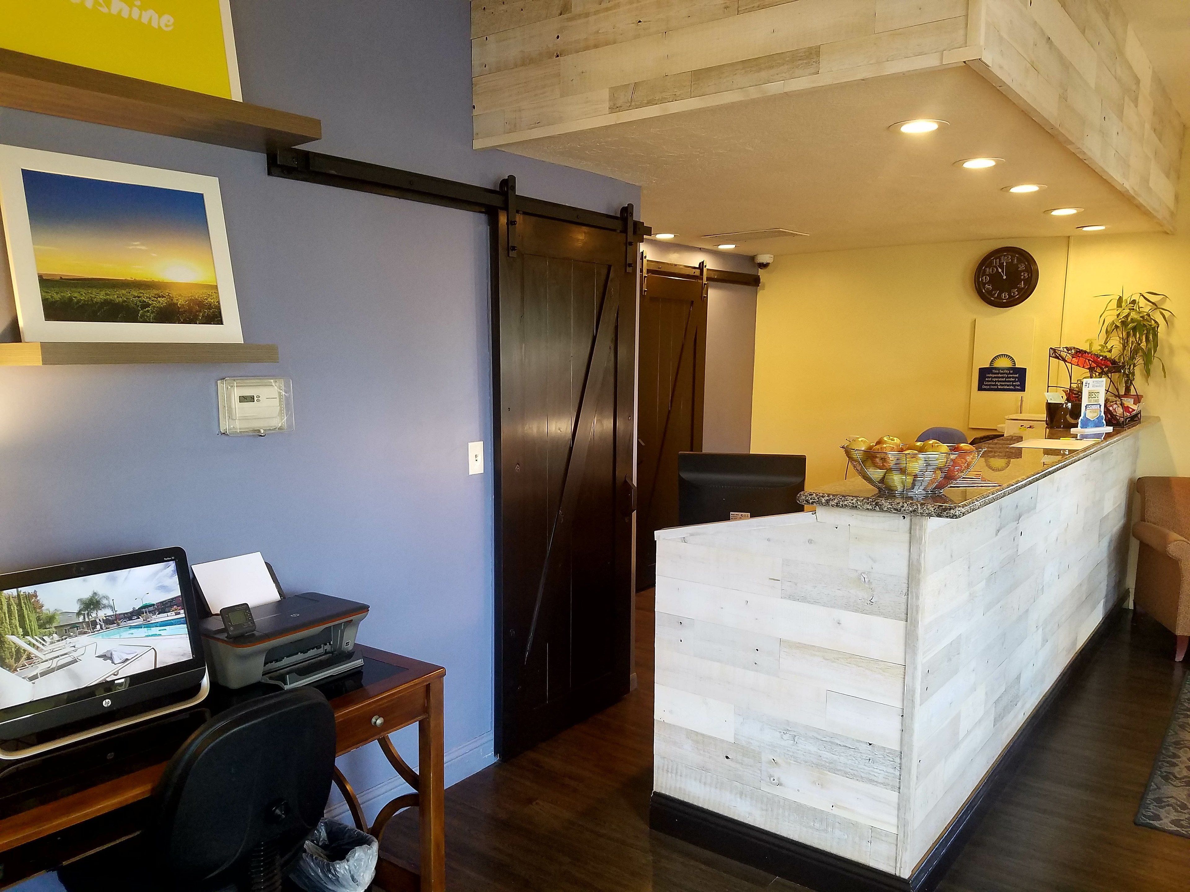 A reception area with a wooden desk, a computer, and a monitor displaying a pool view. There is a sliding door leading to another room, a clock on the wall, and a plant on the reception desk.