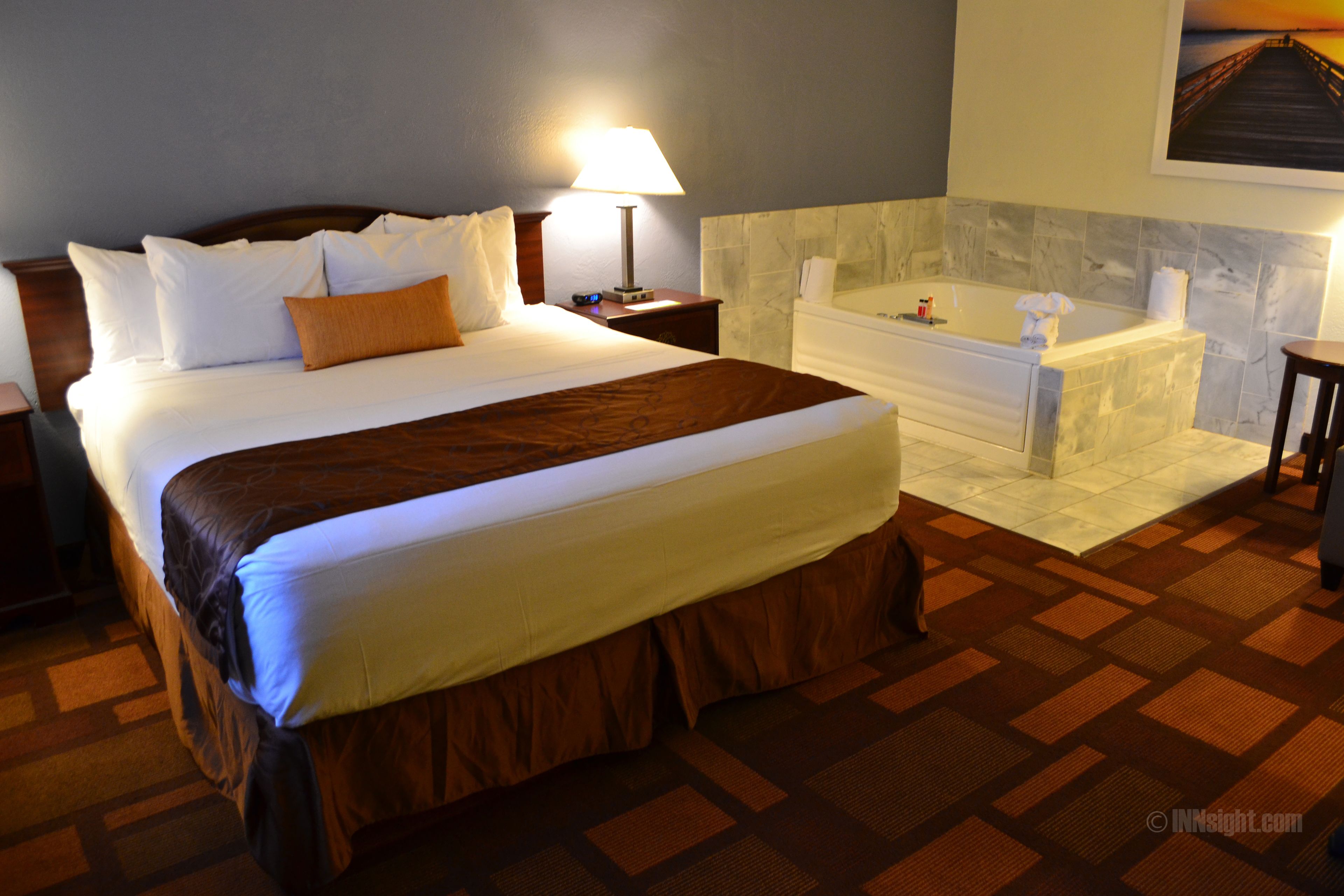 Habitación de hotel con una cama grande, un baño con bañera y una mesita de noche con lámpara.