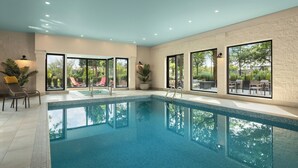 Uma grande piscina interior com água azul cristalina, rodeada por chão e paredes revestidas de azulejos. Há cadeiras e mesas fora da área da piscina, e grandes janelas oferecem vista para o exterior.