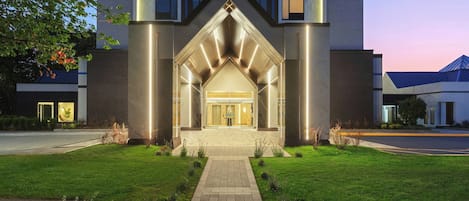 A modern hotel entrance with a well-lit archway, paved pathway, and a grassy lawn.