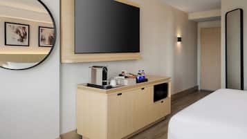 A hotel room with a bed, a TV mounted on the wall, a coffee machine, and a small cabinet with a mirror.