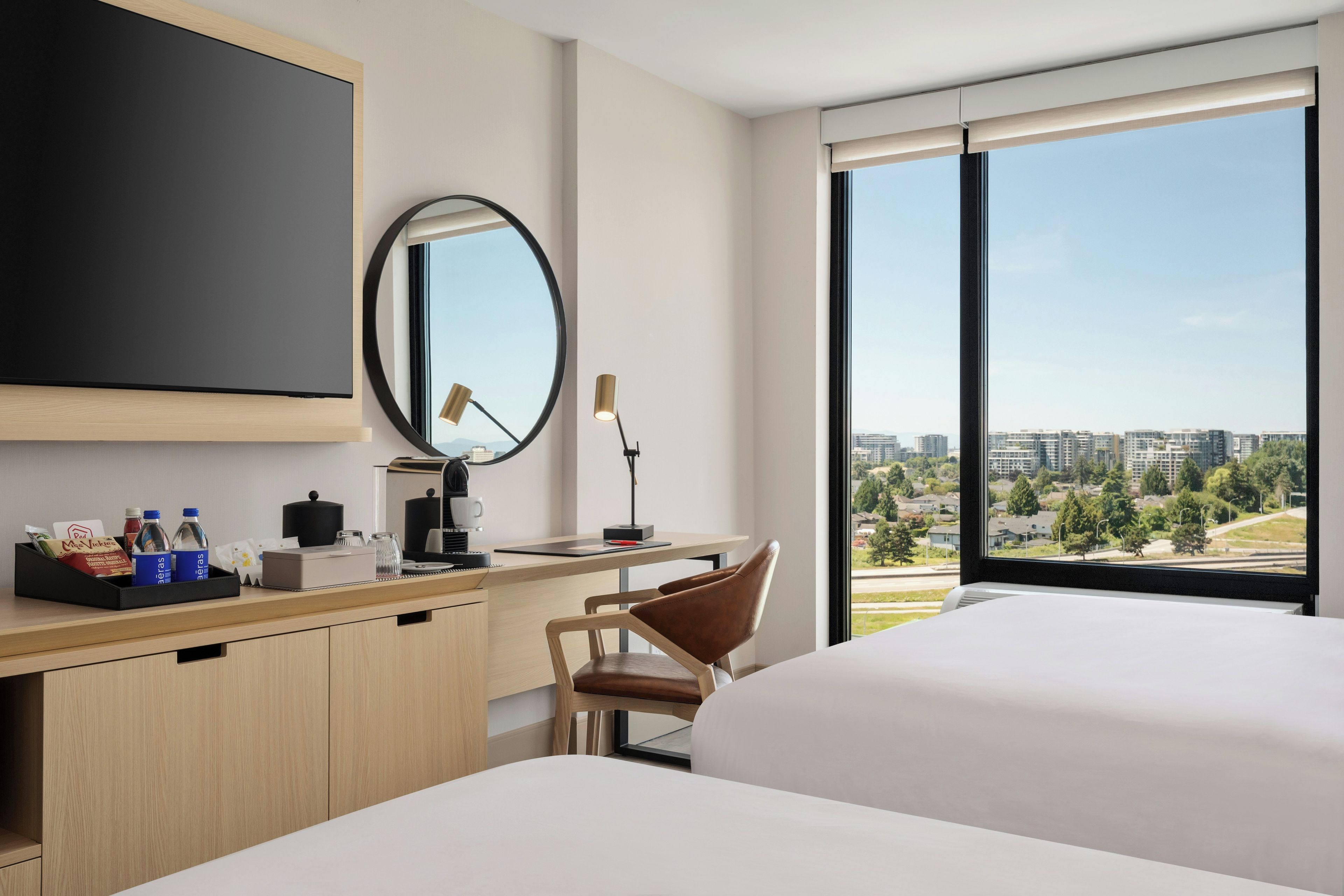 A hotel room with a large bed, a desk with a chair, a flat-screen TV, a mirror, and a window with a city view.