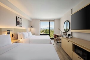 Habitación de hotel con dos camas, un escritorio, televisión y una ventana grande con vista a la ciudad.