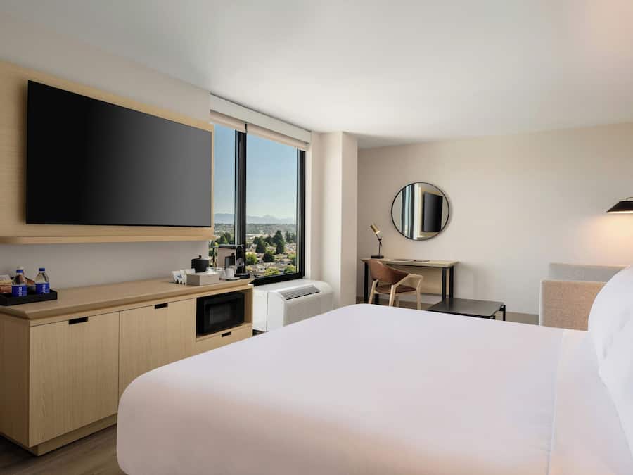 A hotel room with a bed, a desk, a chair, a large window with a view, a flat-screen TV, and a mirror.