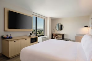 Quarto de hotel com cama, escrivaninha, cadeira, janela grande com vista, televisão de tela plana e espelho.