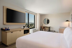 Habitación de hotel con cama, escritorio, silla, ventana grande con vistas, televisor de pantalla plana y espejo.