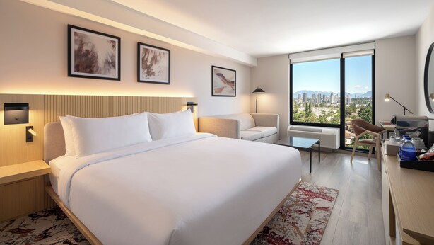 Quarto de hotel moderno com uma cama grande, uma escrivaninha, uma cadeira e vista para a cidade.