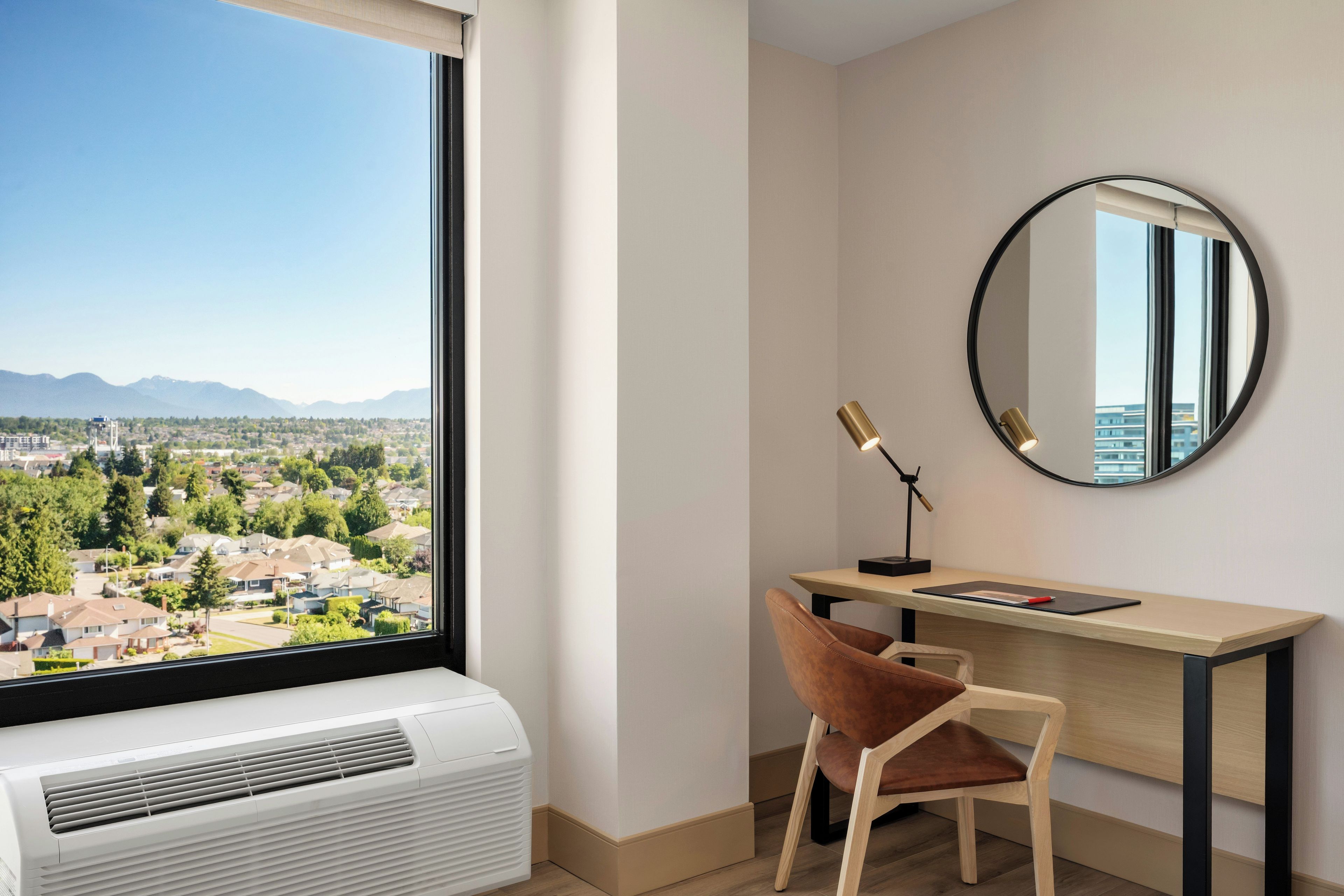 Une chambre moderne dotée d’une grande fenêtre, d’un bureau, d’une chaise, d’un miroir rond et d’un appareil de climatisation.