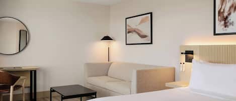 Habitación de hotel con cama, sofá, escritorio, silla, mesita, lámpara y dos cuadros en la pared.