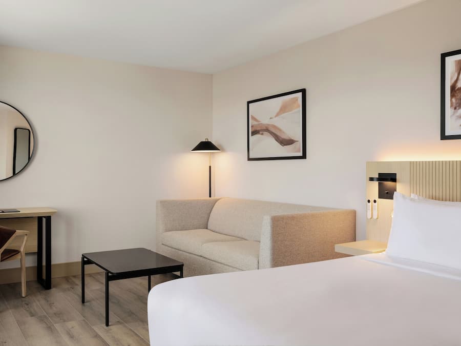 Habitación de hotel con cama, sofá, escritorio, silla, mesita, lámpara y dos cuadros en la pared.