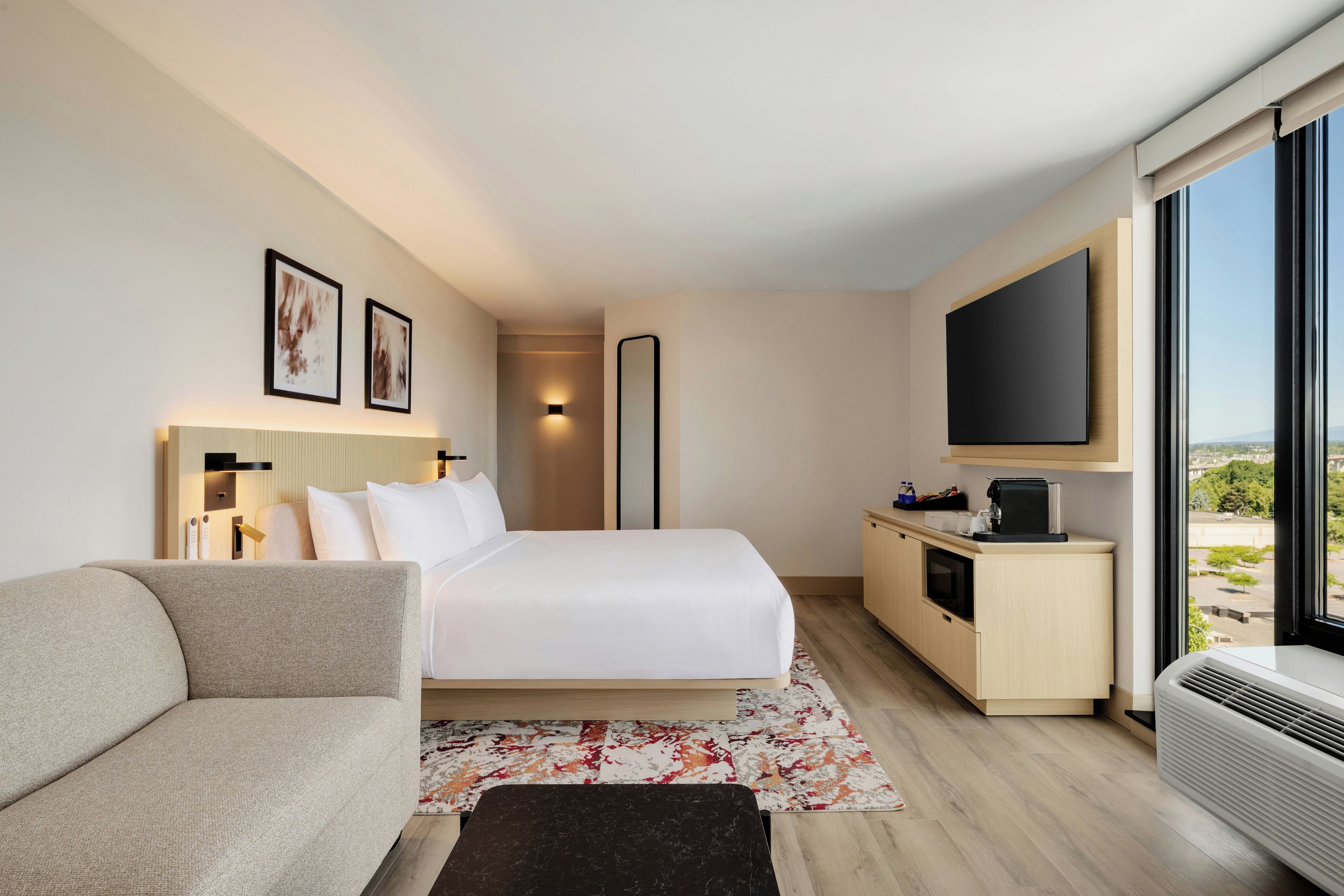 A modern hotel room with a bed, a sofa, a flat-screen TV, a minibar, and a large window offering a city view.