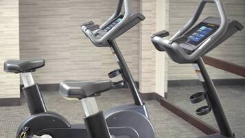 Two stationary bikes with digital interfaces, likely in a gym setting with a tiled wall in the background.