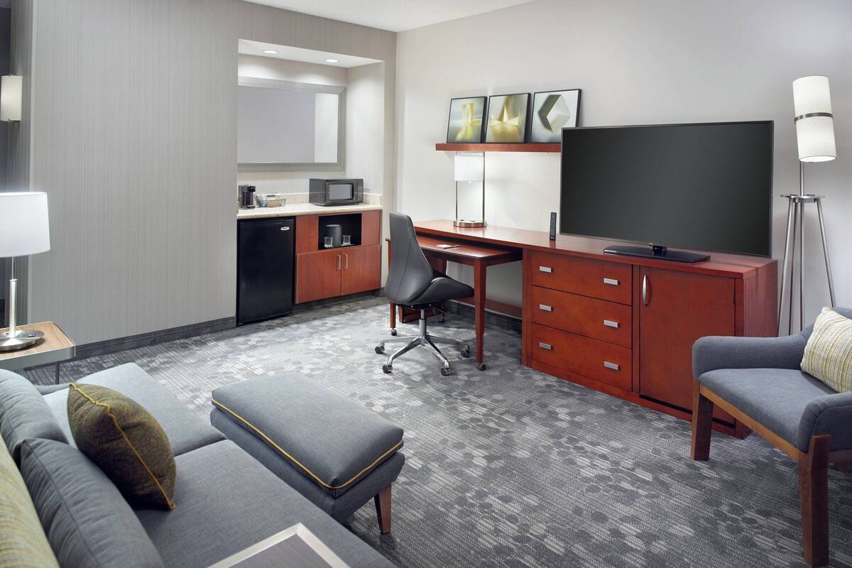 Une chambre d’hôtel avec un canapé gris, un petit pouf, un bureau avec une chaise, une télévision, un four à micro-ondes, une machine à café et une lampe.