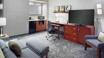 A hotel room with a gray sofa, a small ottoman, a desk with a chair, a television, a microwave, a coffee maker, and a lamp.