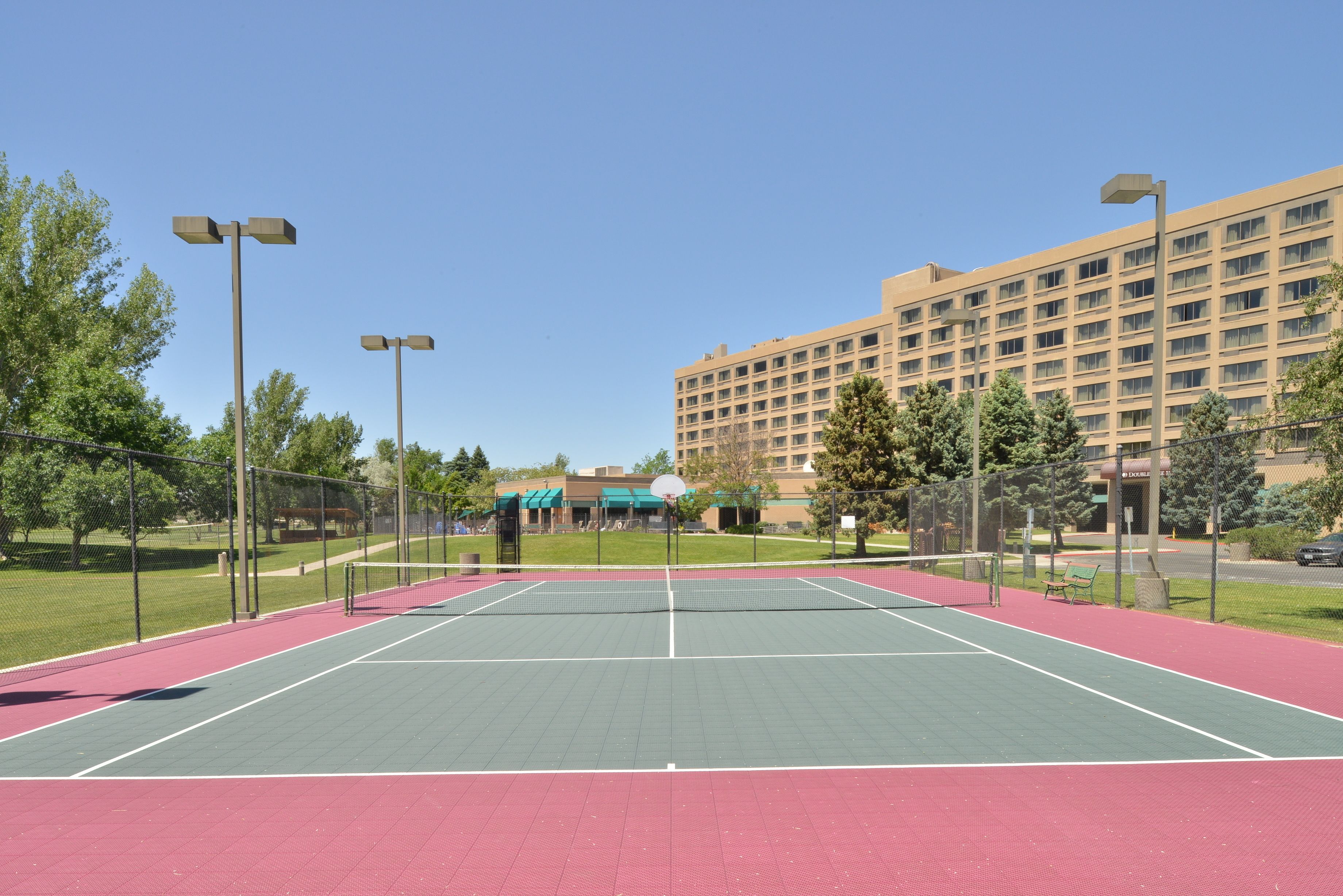 Ein Außen-Tennisplatz mit Netz, umgeben von einem Zaun, und ein großes Gebäude im Hintergrund.