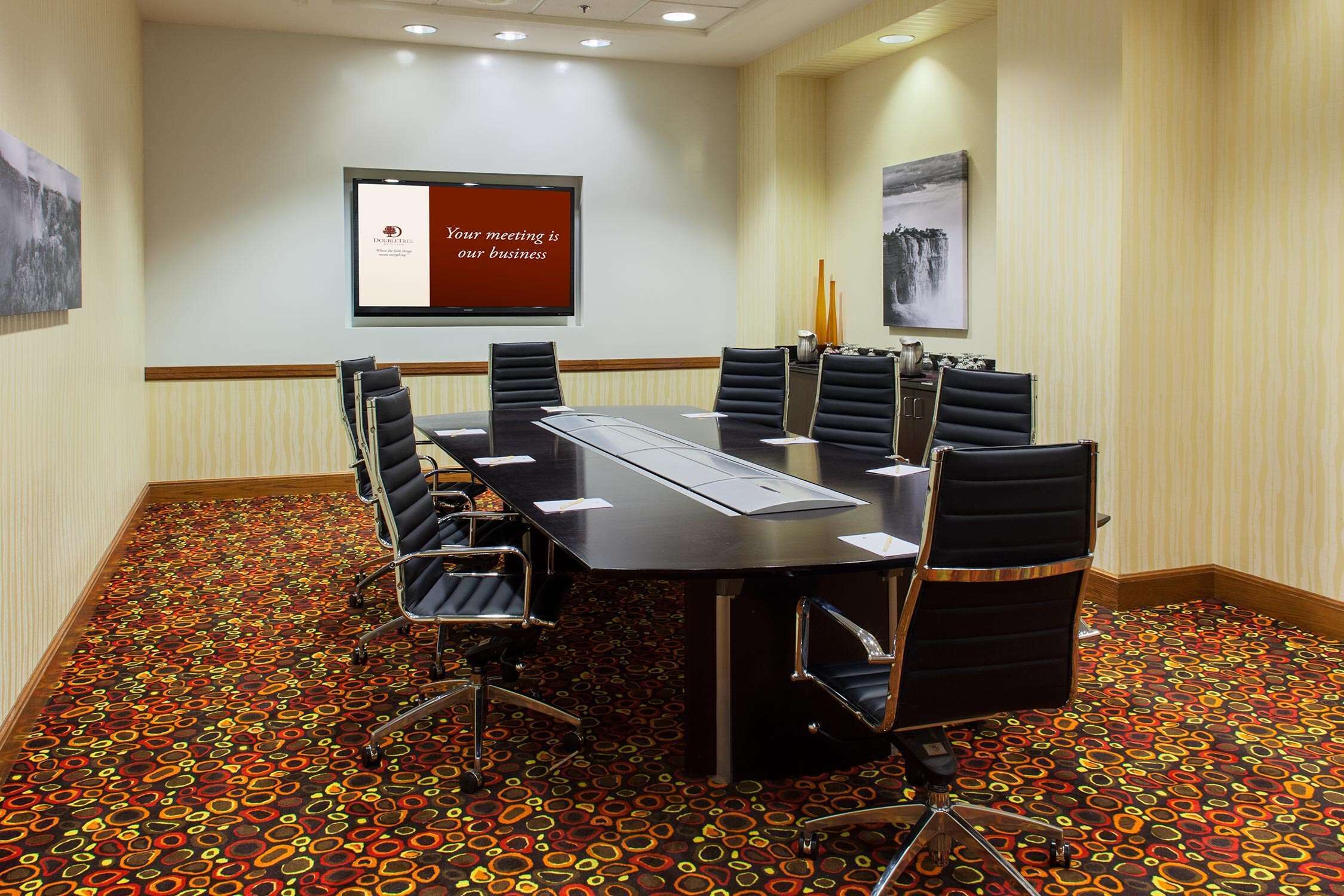 A conference room with a long table, six chairs, a projector screen, and patterned carpet.