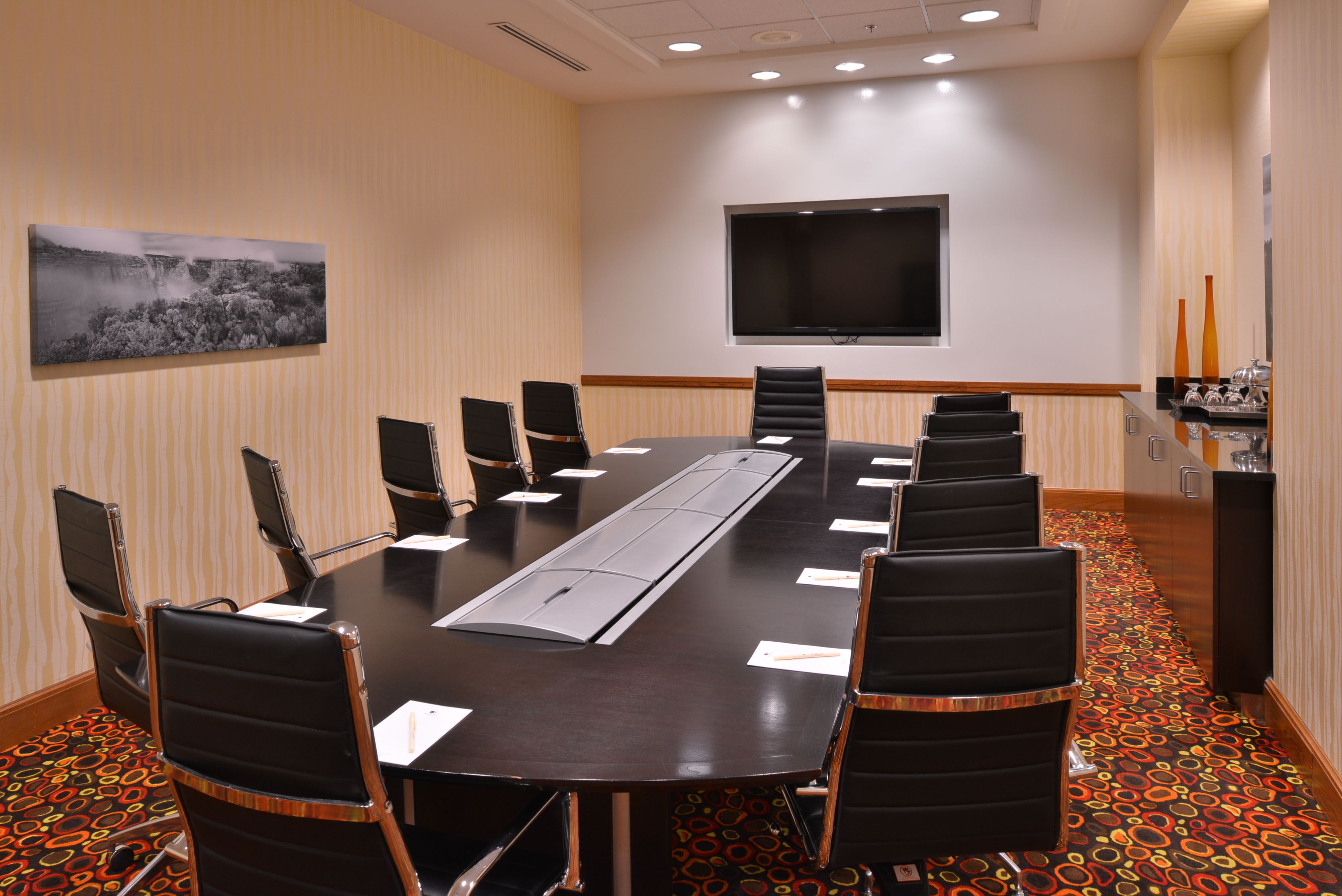 A conference room with a long table, chairs, a flat-screen TV, and a decorative wall piece.