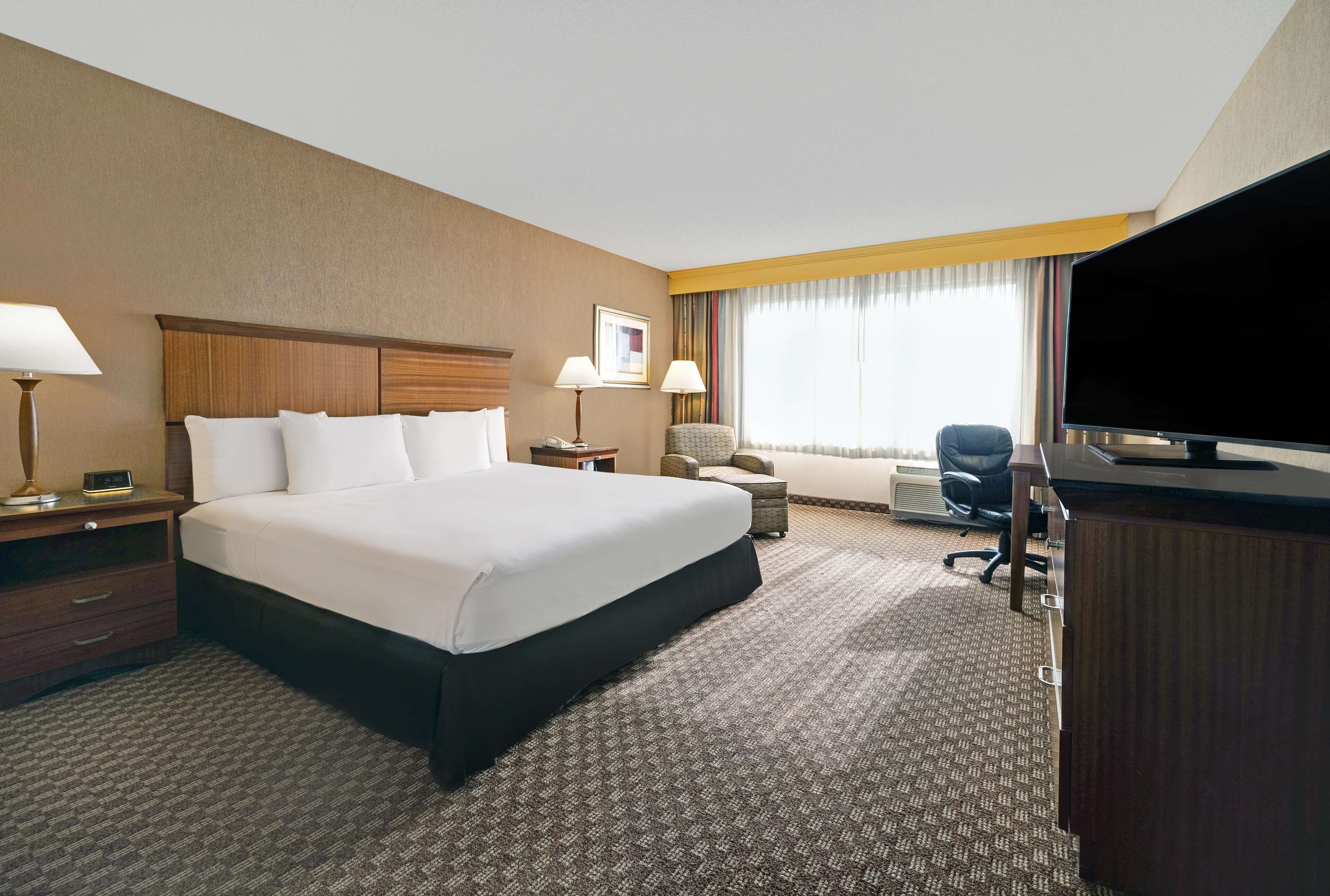 Une chambre d’hôtel avec un grand lit, un bureau avec un ordinateur, une chaise, une télévision et une fenêtre avec des rideaux.