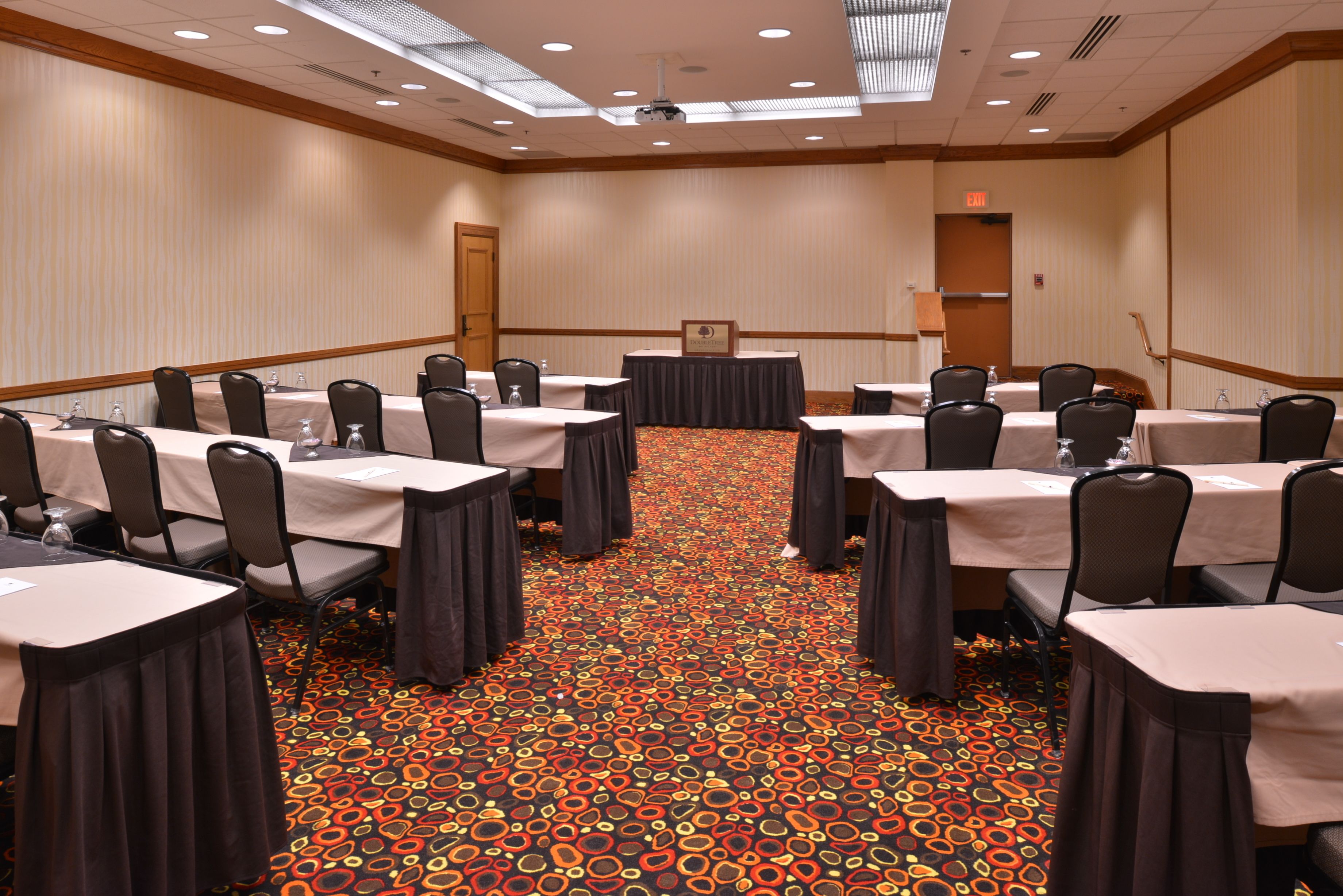 Une salle de conférence équipée de tables et de chaises, recouverte d’un tapis à motifs et dotée d’un écran de projection.