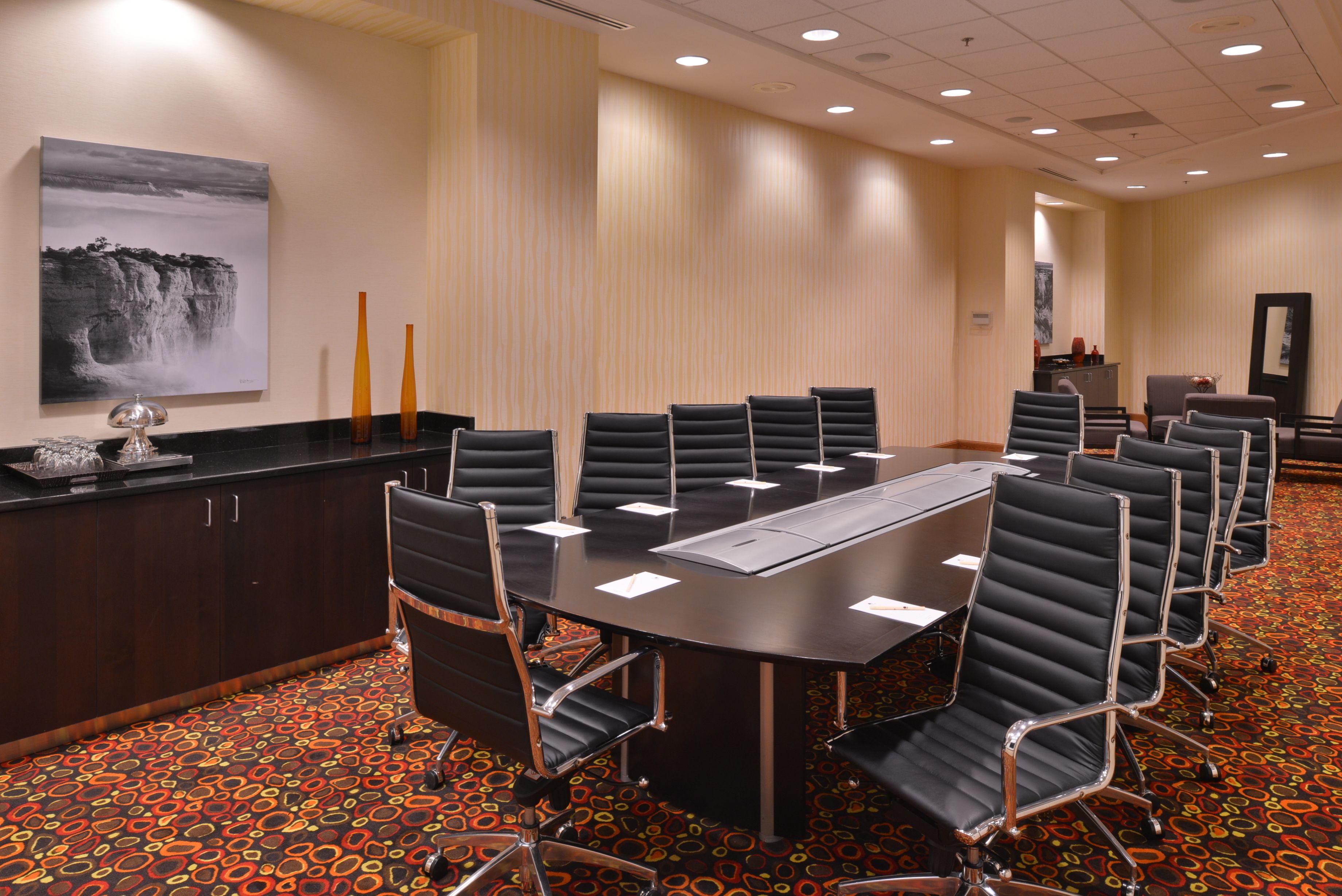 Une salle de conférence avec une grande table, plusieurs chaises, un comptoir orné d’un vase et un tableau accroché au mur.