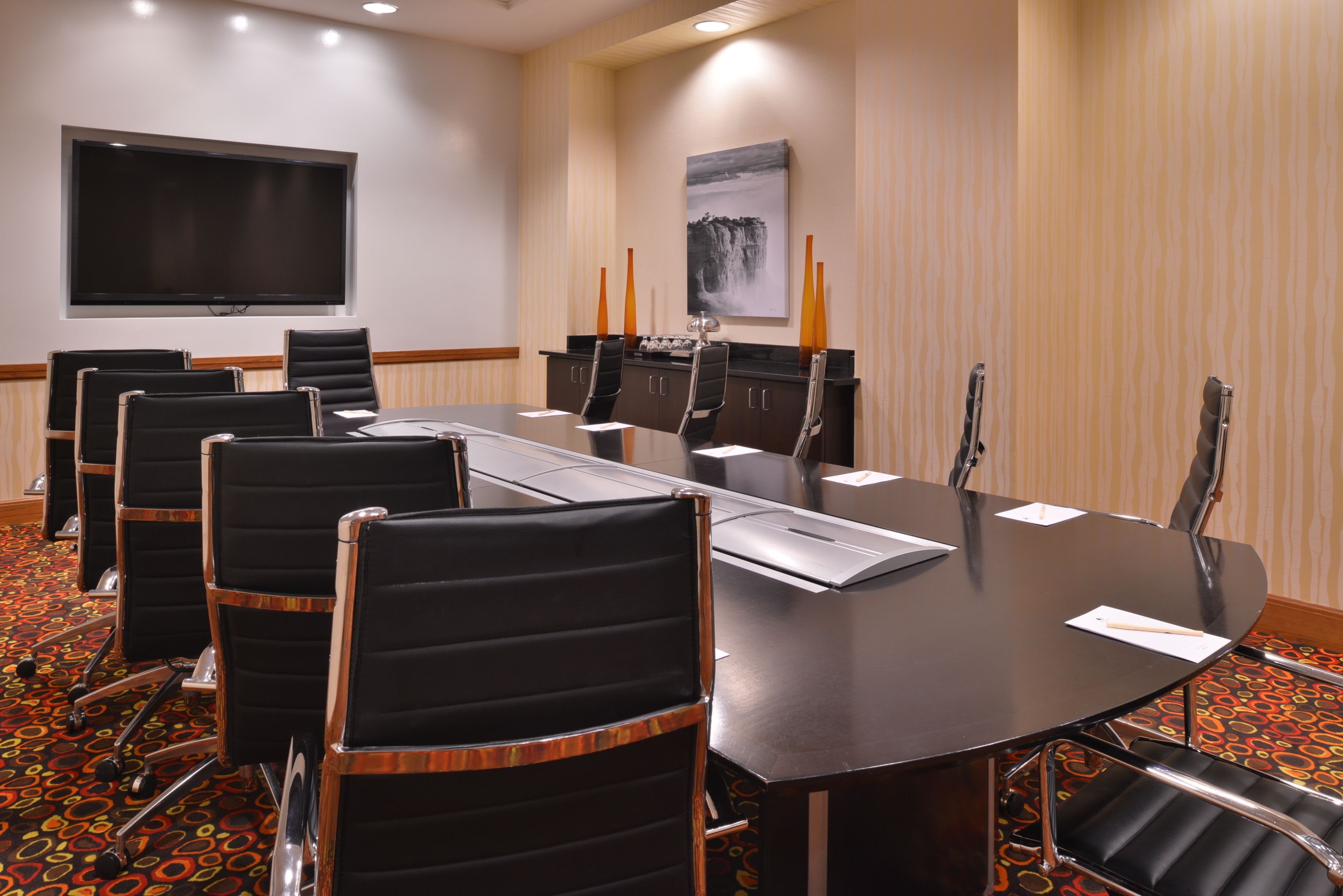 Un salón de conferencias con una gran mesa, sillas negras, un televisor de pantalla plana y un cuadro en la pared.