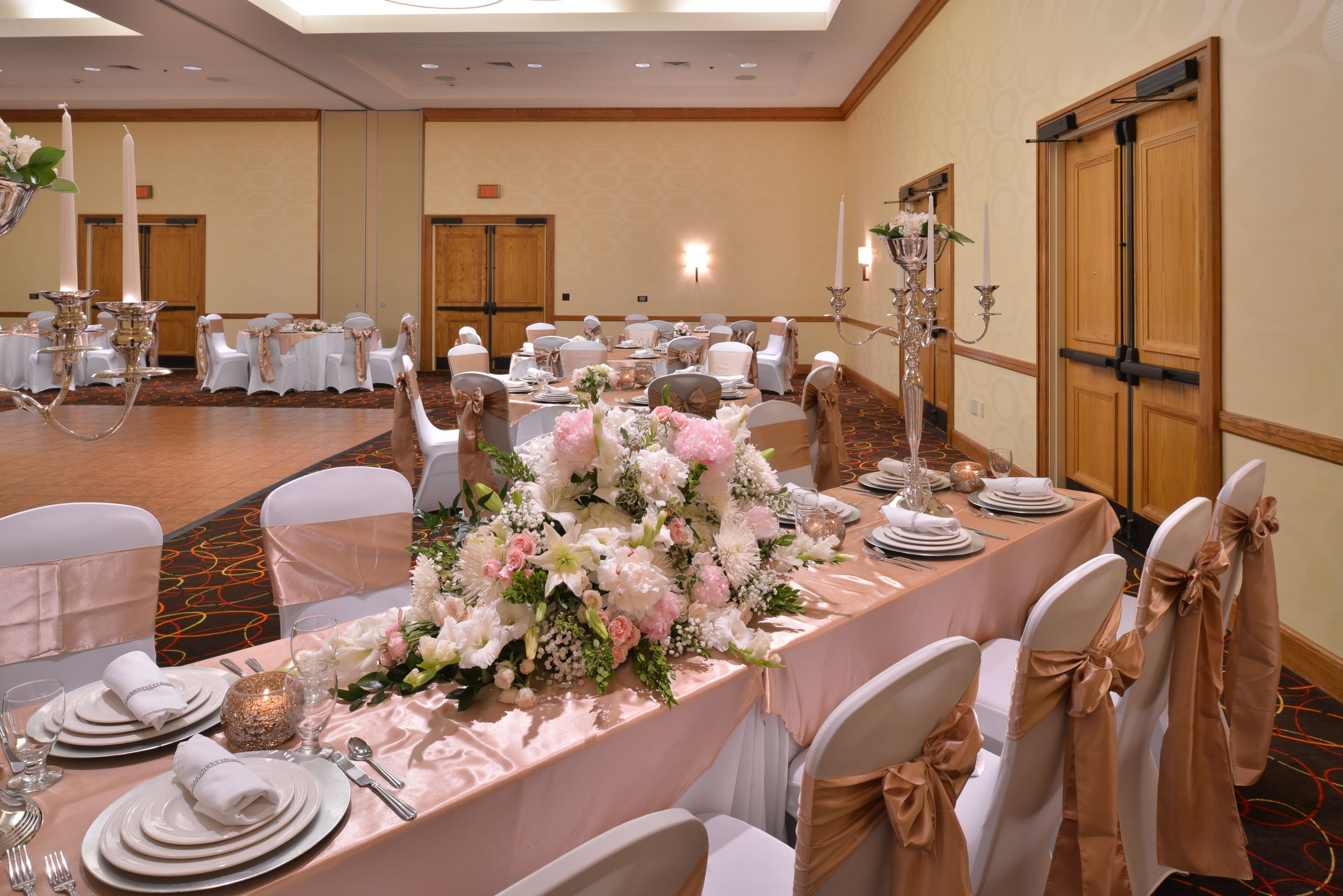 A banquet hall with round tables set for a formal event, featuring white chairs with gold sashes, floral centerpieces, and a chandelier.