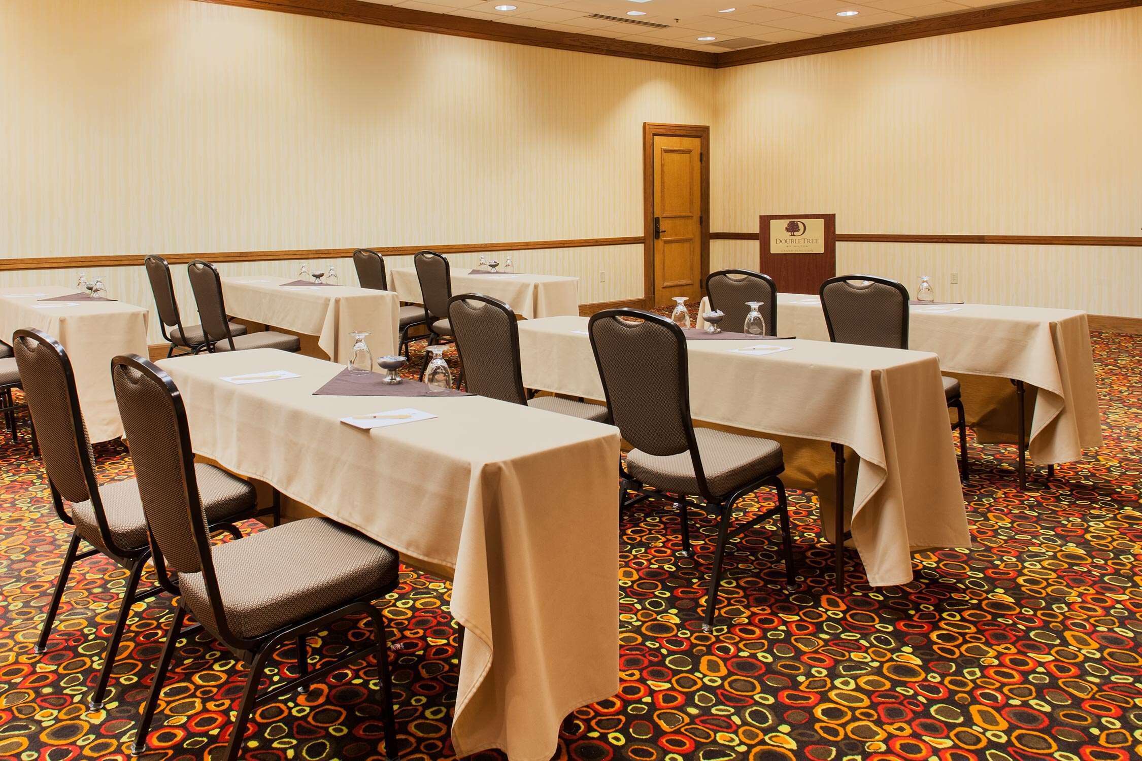 A conference room with multiple tables and chairs, carpeted floor, and a door in the background.