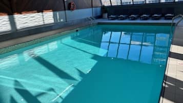 An indoor swimming pool with clear water, surrounded by lounge chairs and a view of a building through large windows.