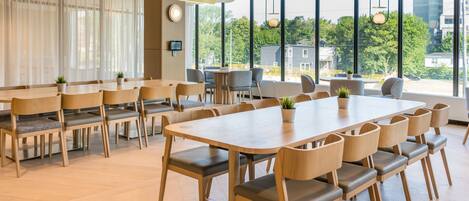 Una sala de conferencias moderna con mesas y sillas de madera, ventanas amplias con vistas a edificios y árboles, e iluminación empotrada en el techo.