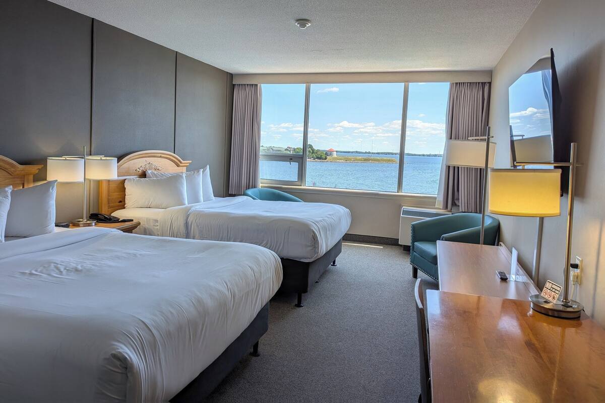 Habitación de hotel con dos camas, un escritorio, una silla y un amplio ventanal con vista al agua.
