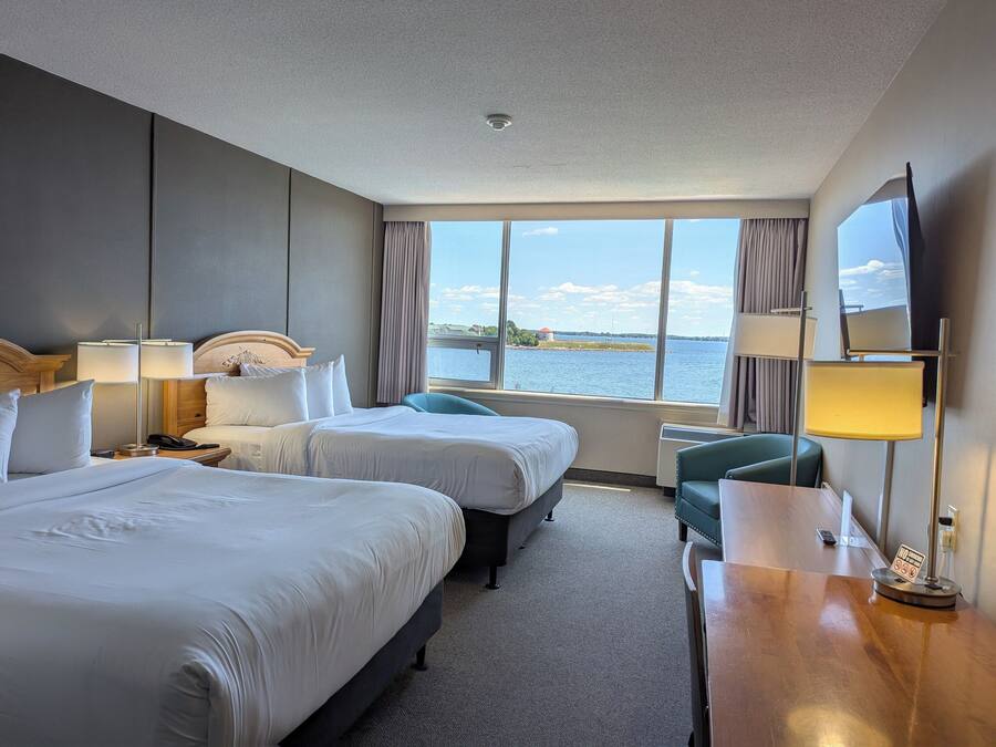 A hotel room with two beds, a desk, a chair, and a large window offering a view of the water.