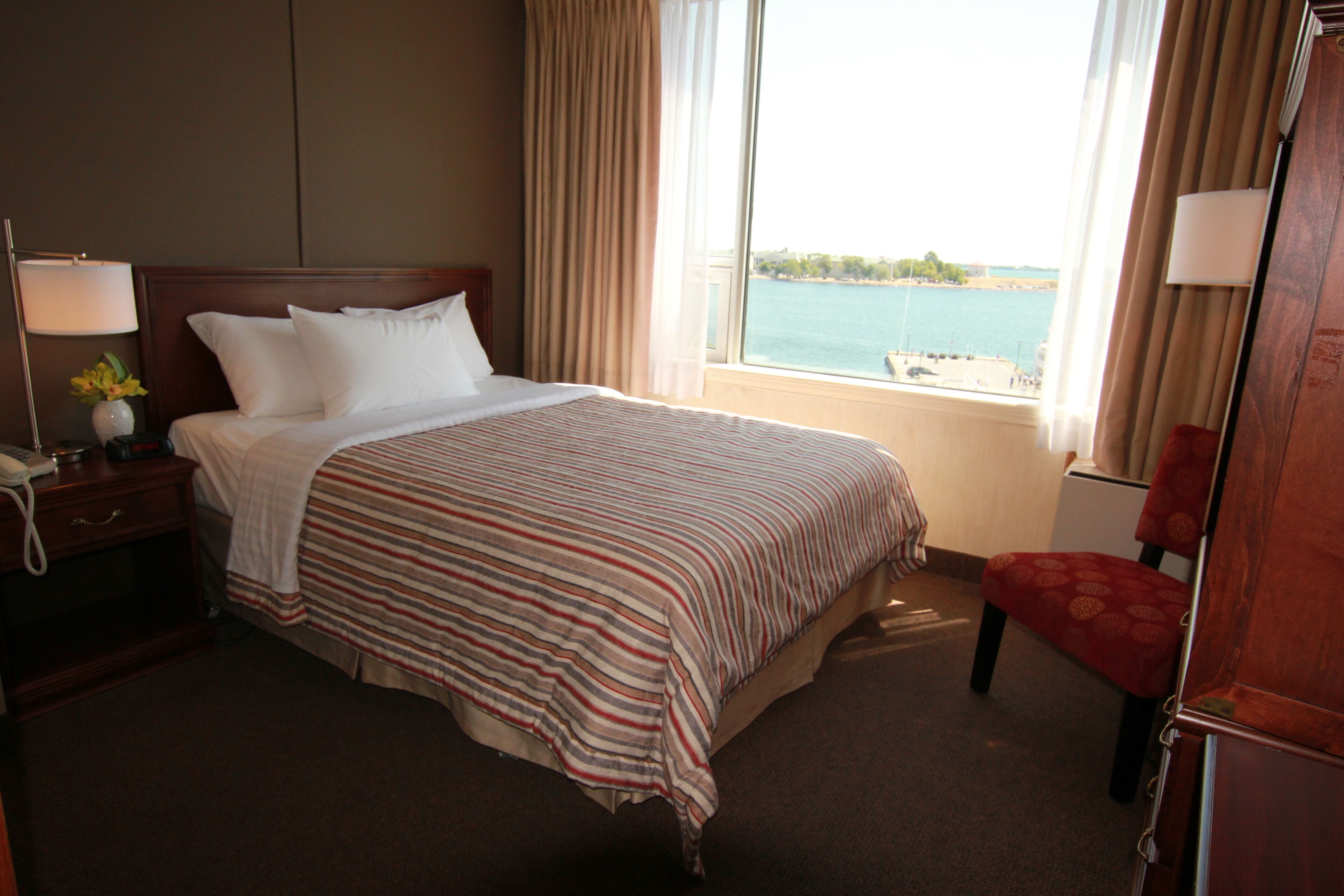Habitación de hotel con una cama grande, una mesita de noche con lámpara, una silla y vista al mar a través de la ventana.