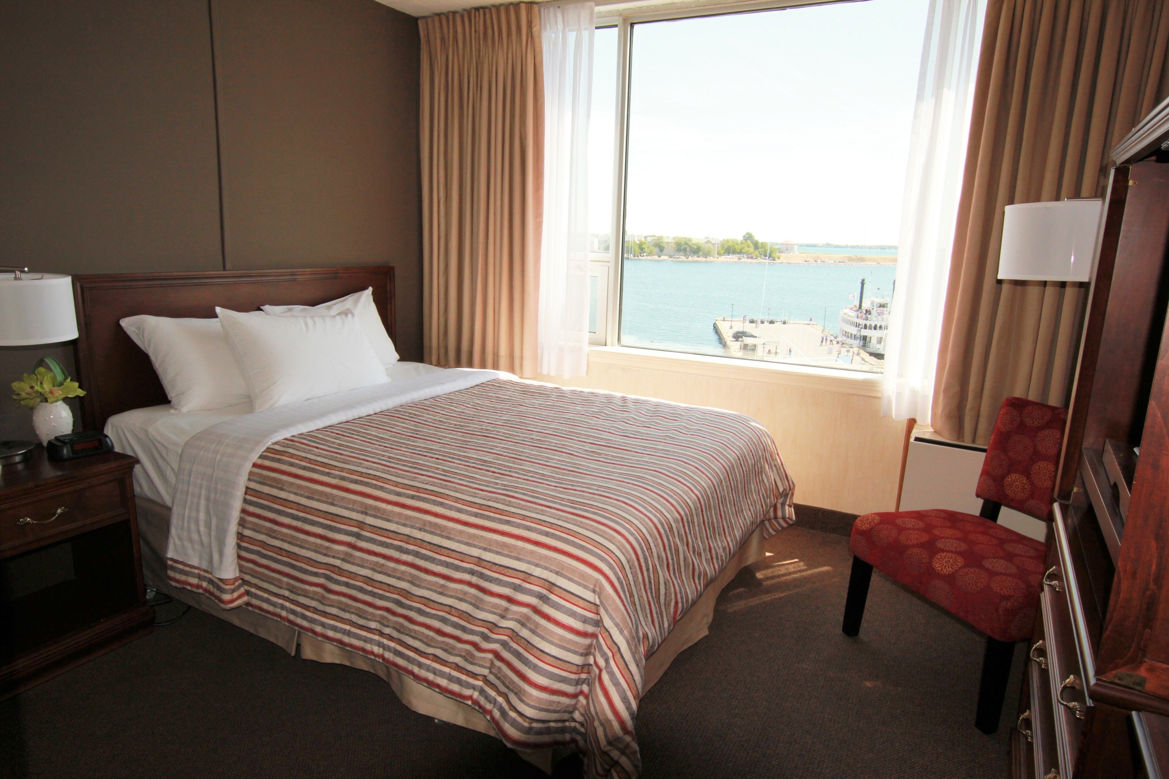Ein Hotelzimmer mit Bett, Sessel, Nachttisch, Lampe und Blick auf ein Gewässer durch das Fenster.