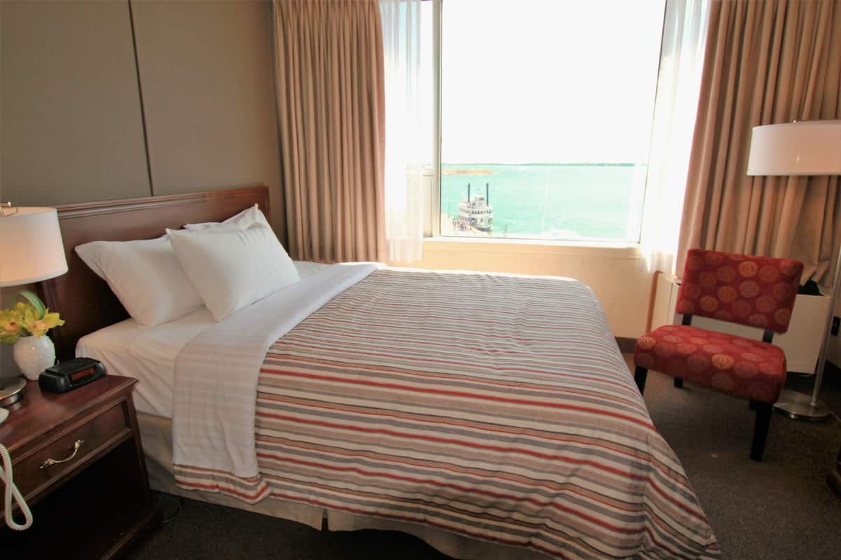 Habitación de hotel con cama, mesita de noche con lámpara, una silla y ventana con vista al mar.