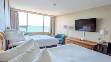 A hotel room with two beds, a desk, a TV, and a view of the sea.
