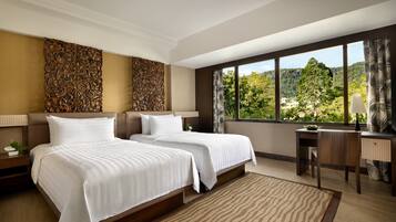 A hotel room with two beds, a desk, and a large window offering a view of a forested area.