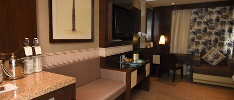 A hotel room with a seating area, a television, a desk with a lamp, and a wall with a palm tree painting.