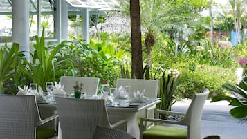 An outdoor dining area with wicker furniture and a glass table, surrounded by lush greenery and a view of a tropical landscape.