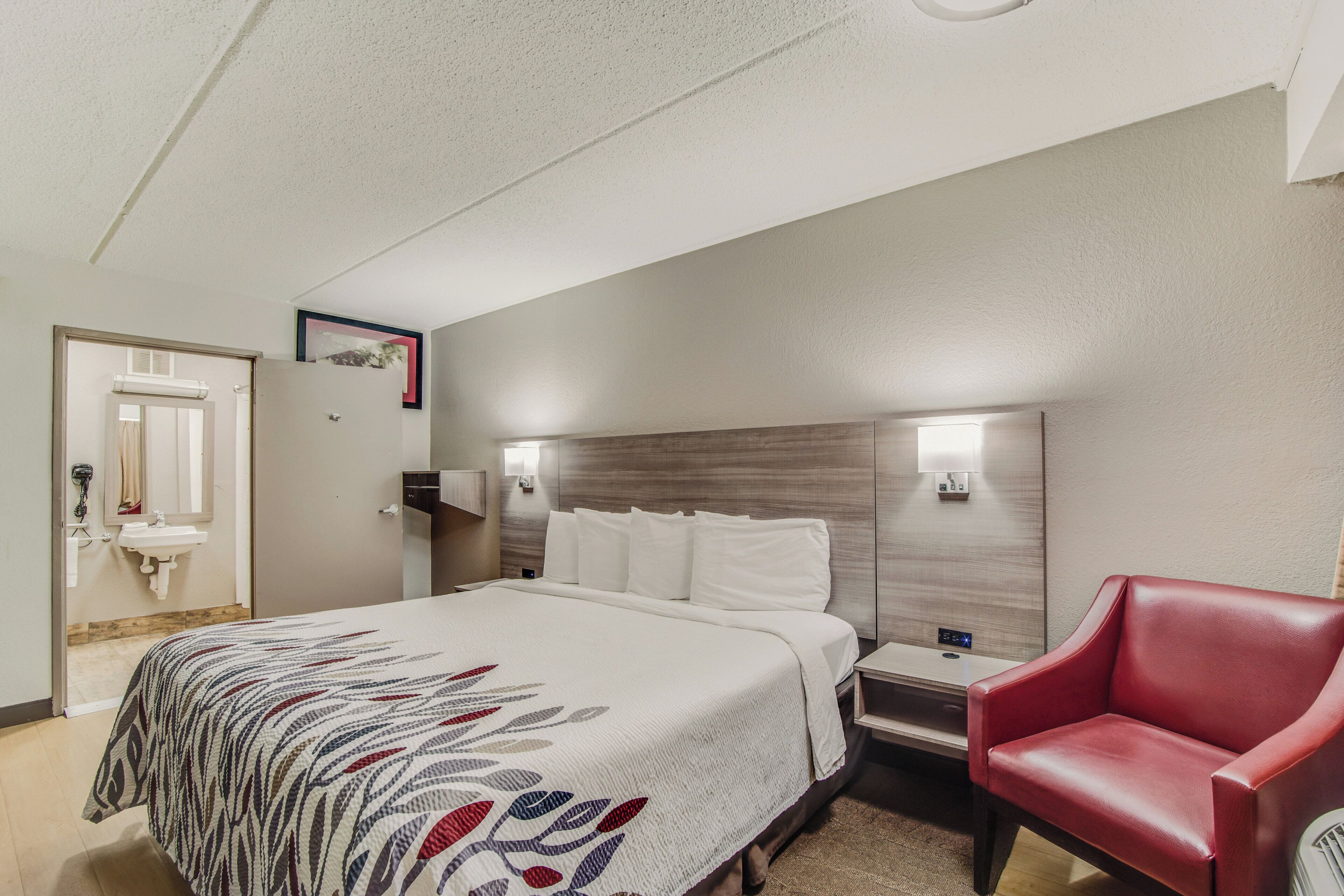 A hotel room with a bed, a red chair, a nightstand, a wall-mounted lamp, and a door with a mirror and a hairdryer.