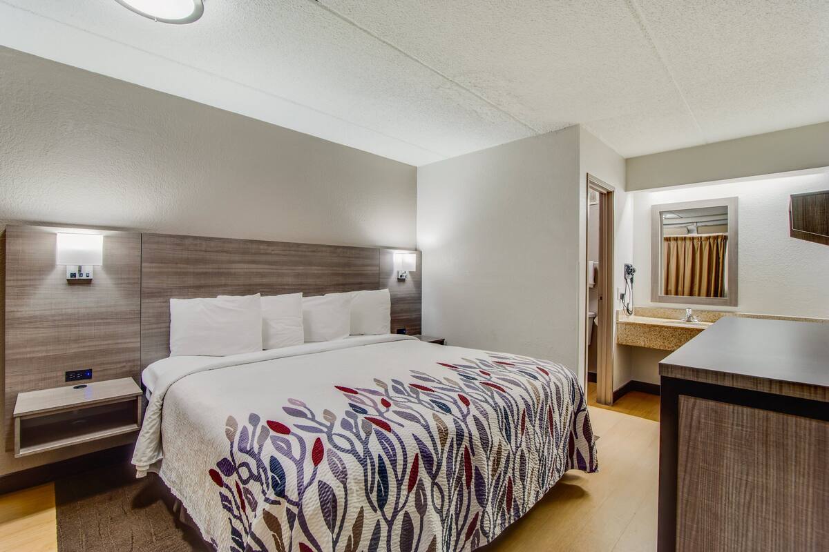 A hotel room with a bed, bedside tables, a nightstand, a mirror, and a bathroom with a sink and shower.