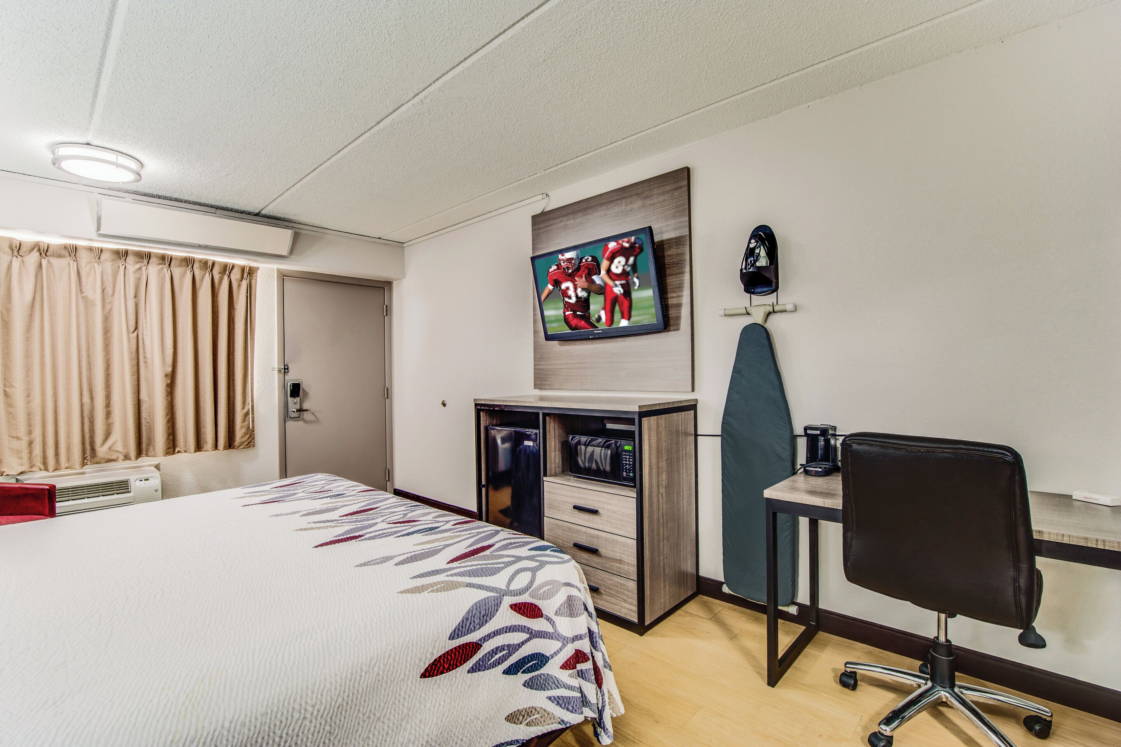 A hotel room with a bed, a desk with a computer, a chair, a microwave, and a television on the wall.