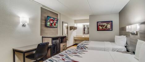 A hotel room with two beds, a desk, a chair, a refrigerator, a microwave, and a television.