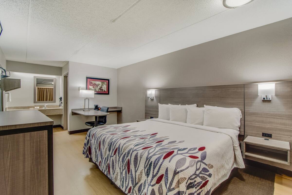A hotel room with a bed, bedside table, desk, and a bathroom with a mirror and towel rack.