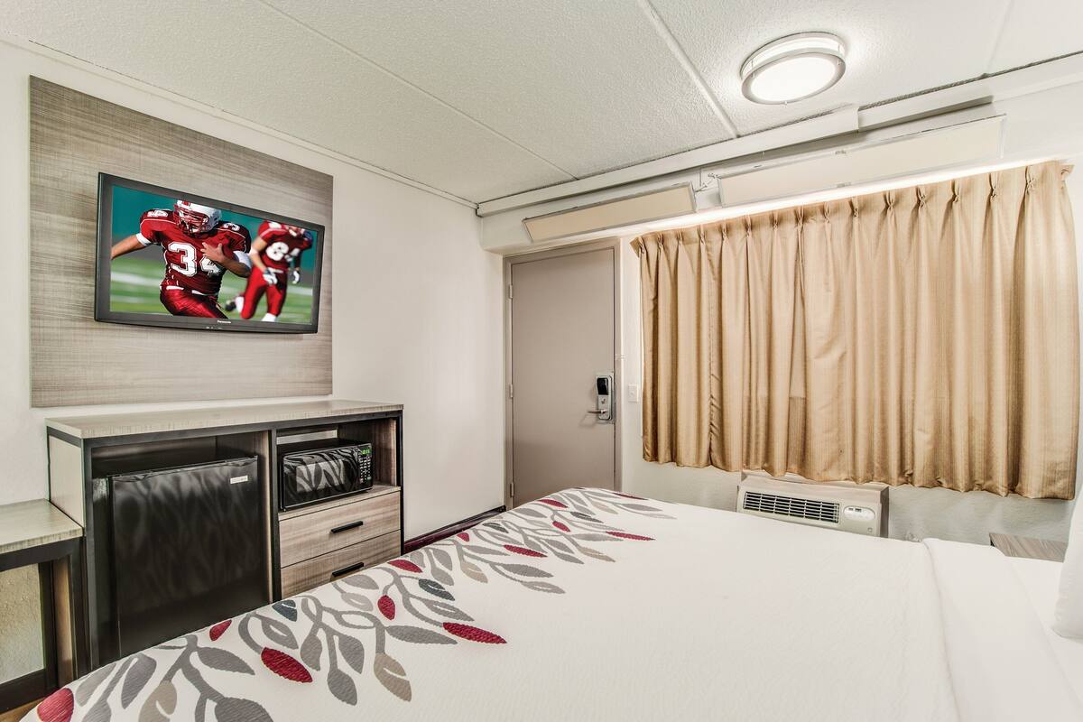 Habitación de hotel con cama, televisor montado en la pared, minibar y ventana con cortinas.