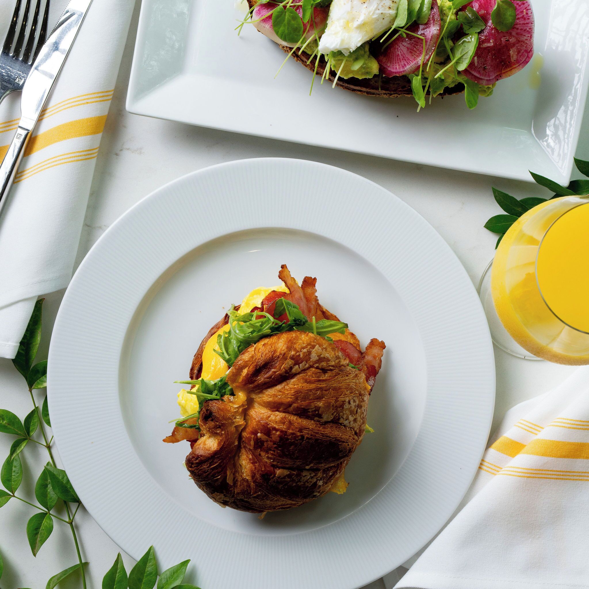 Un plato de desayuno con un croissant, un huevo, vegetales verdes y un vaso de jugo de naranja sobre un mantel blanco.