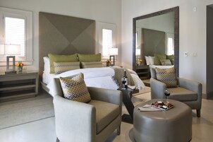 Habitación de hotel moderna con una cama grande, dos sillones, un taburete redondo y una mesa de comedor con una copa de vino y fruta.