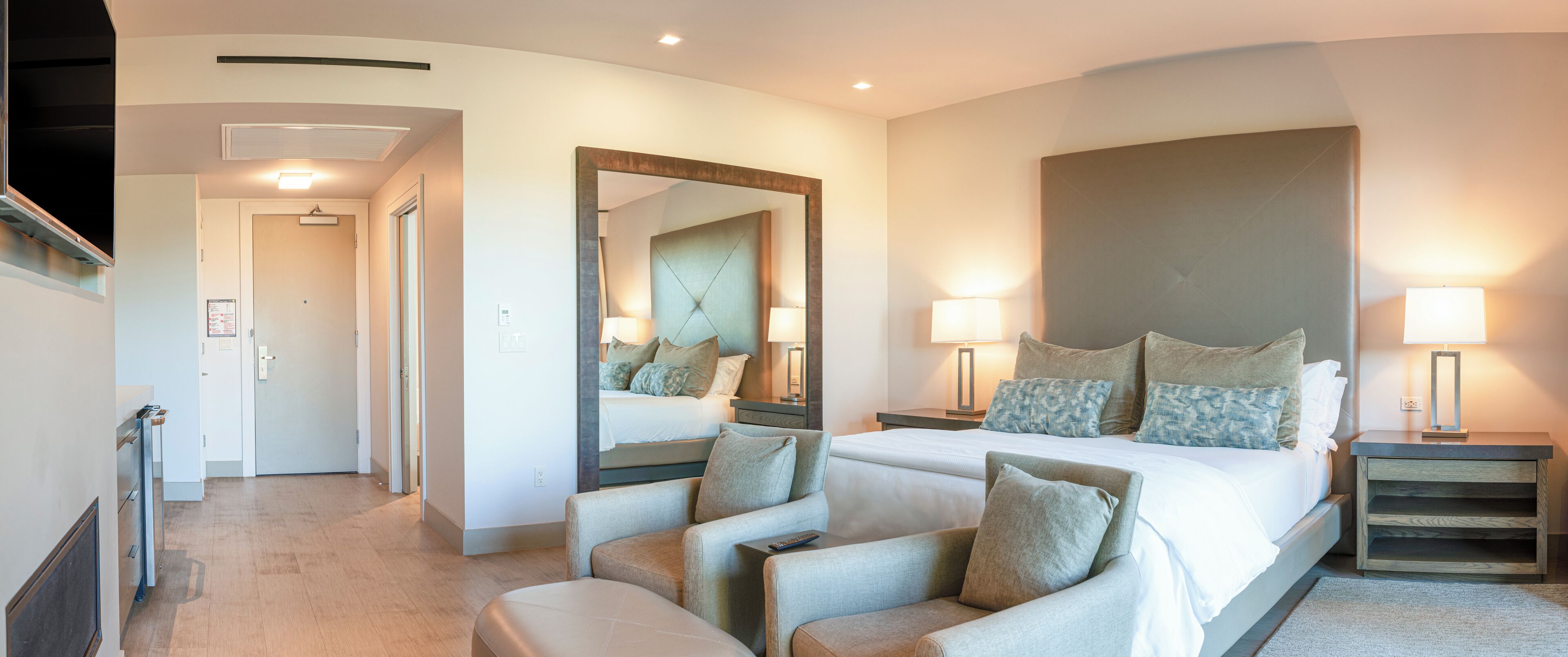 A hotel room with a large bed, two bedside tables with lamps, a seating area with chairs, and a large mirror.