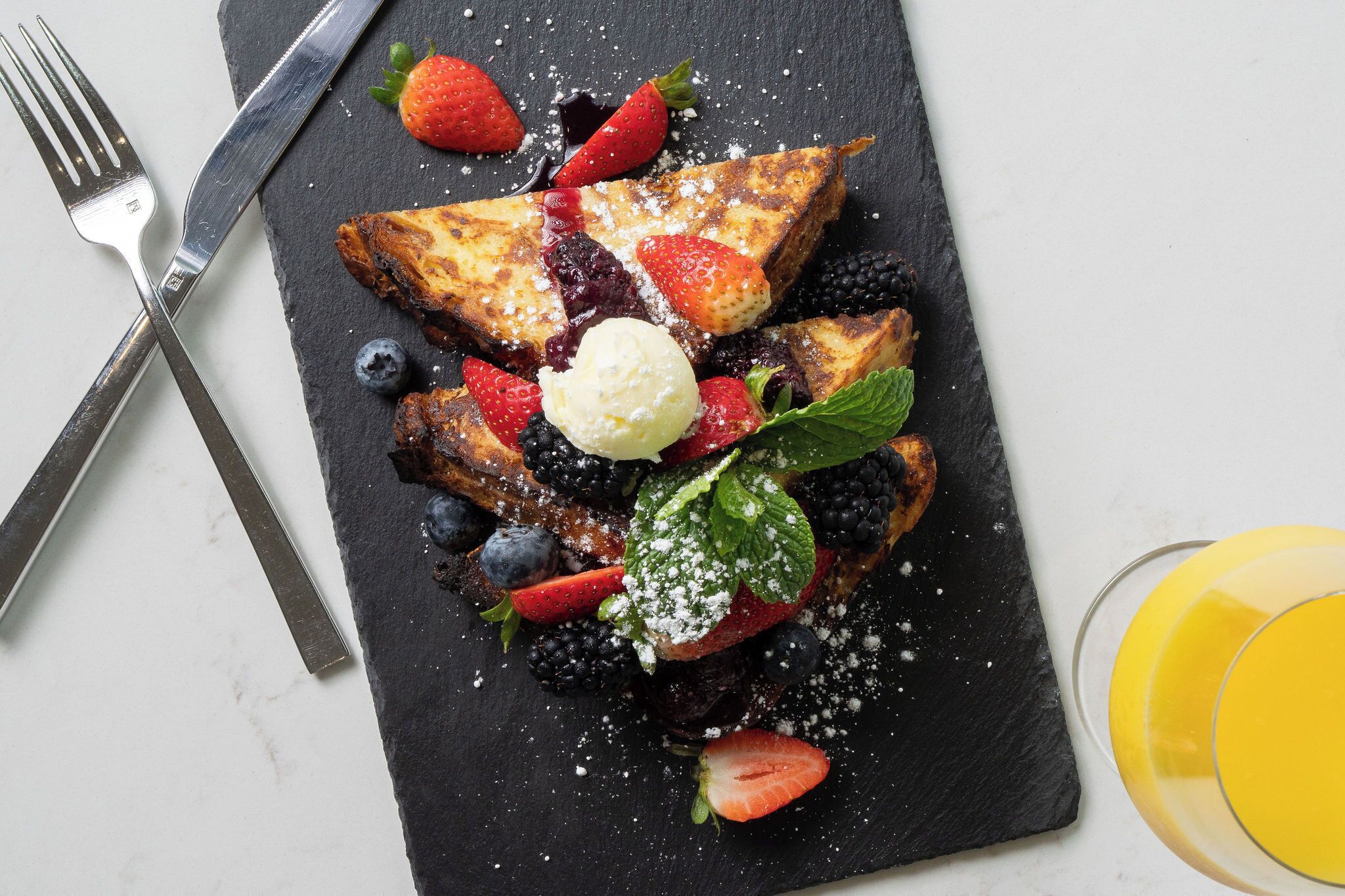 Une assiette de petit-déjeuner avec des tranches de pain perdu, des fraises, des myrtilles, des mûres, de la menthe et une boule de glace, le tout présenté sur une planche d’ardoise.
