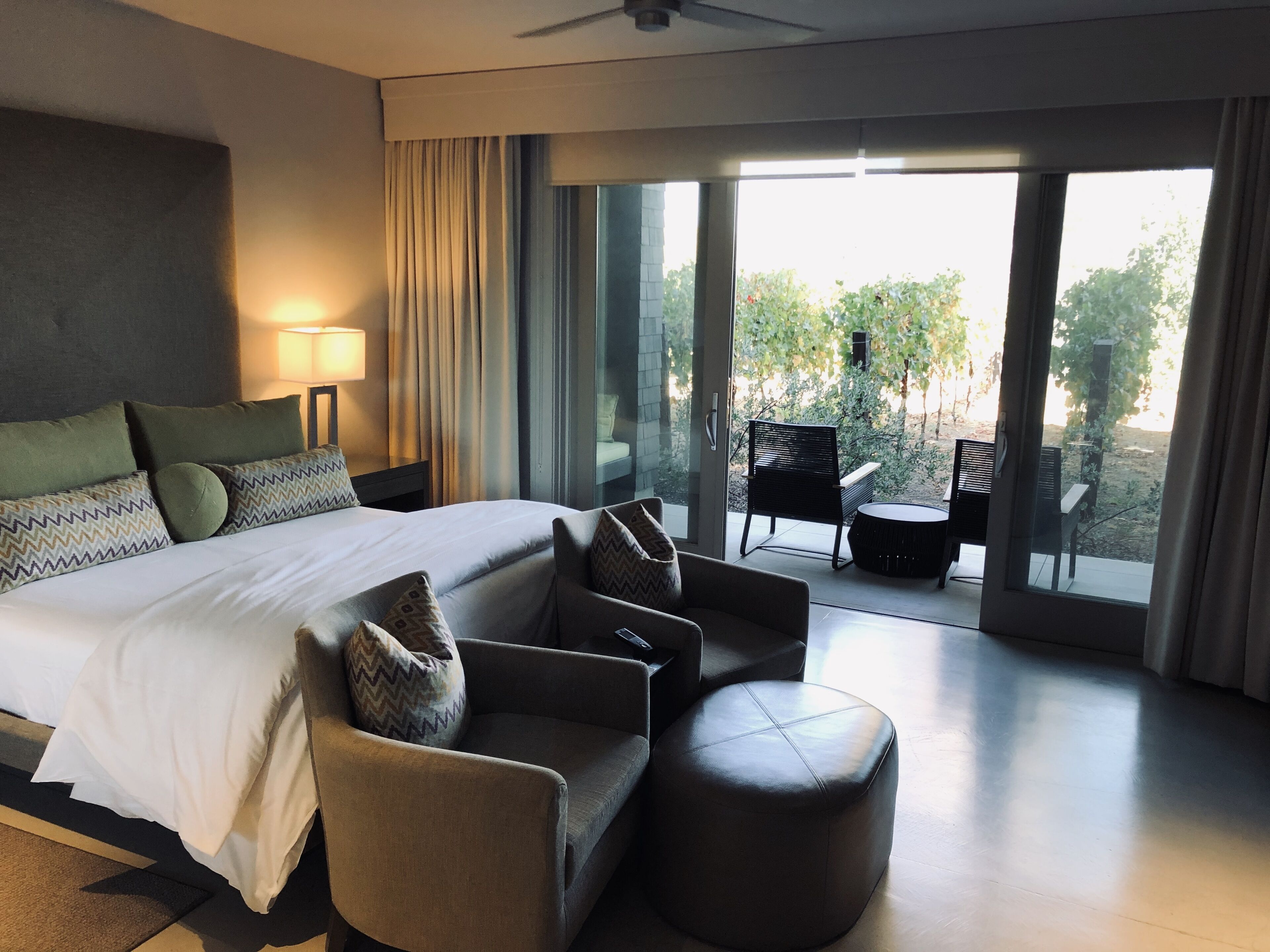 Una habitación de hotel moderna con una cama grande, un sofá, un otoman redondo y un balcón con mobiliario exterior.